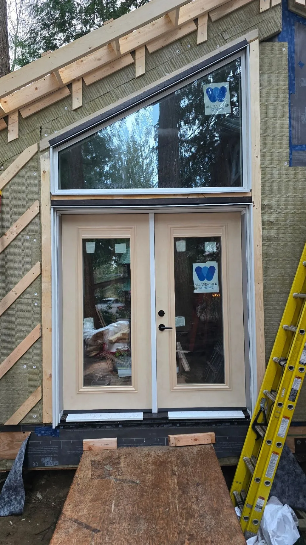 An exterior door with a transom and adjacent window installed in a timber-framed opening, with a ladder nearby.