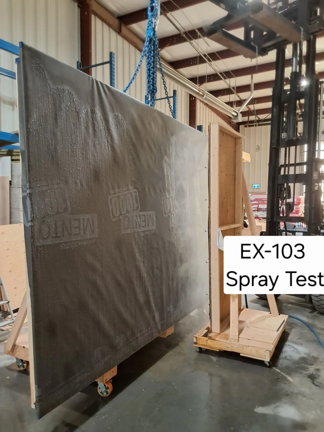 A wall panel on a wheeled stand with water beading across it and a visible EX-103 Spray Test label.
