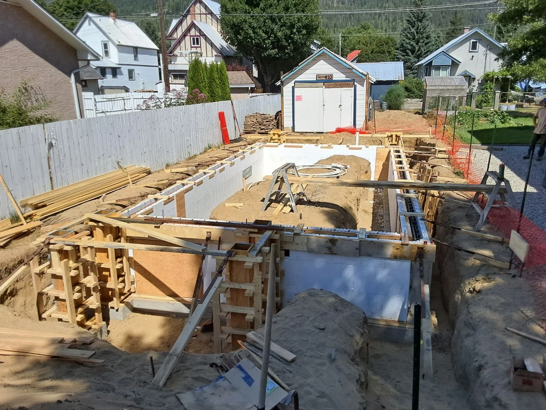 A residential foundation excavation with concrete formwork, gravel, and neighboring homes visible beyond it.
