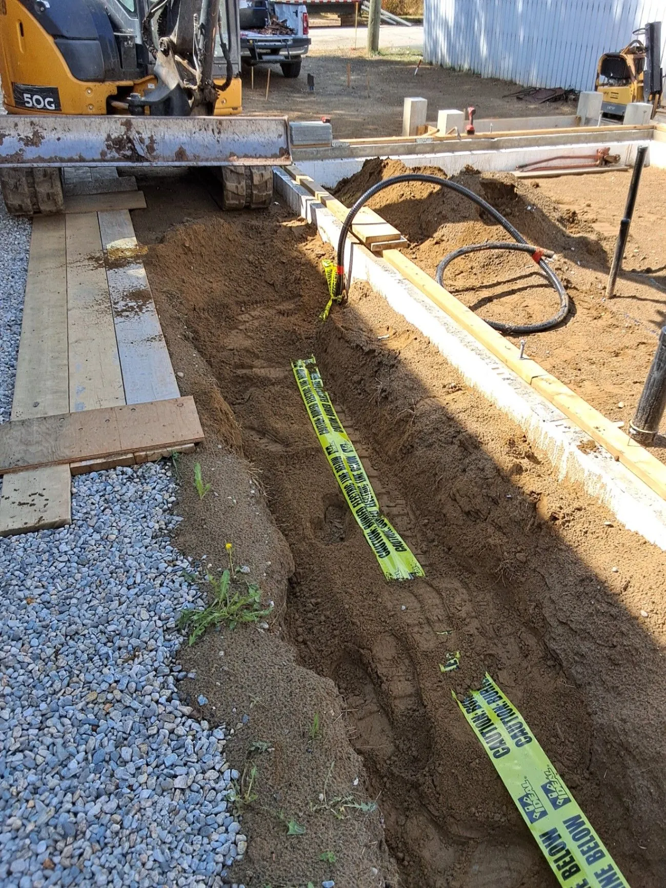 A narrow excavated trench with conduit beside a gravel edge and concrete work.
